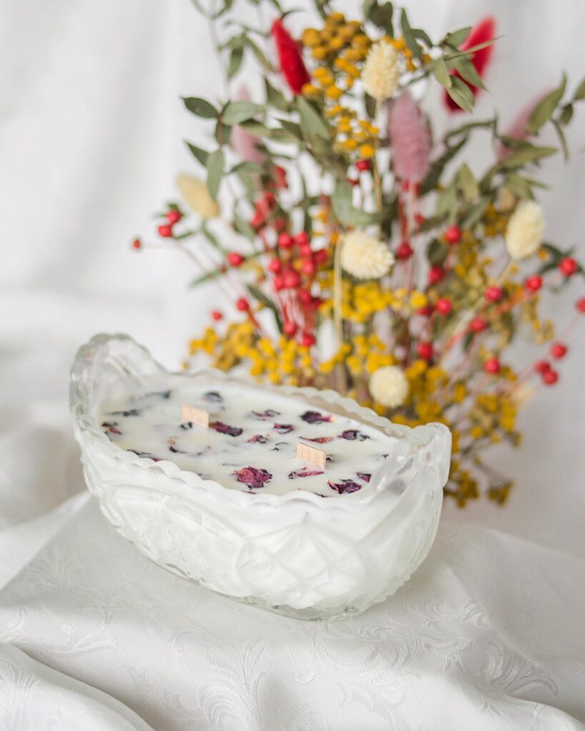 świeca w kryształowym naczynku z płatkami kwiatów, stojąca na białym obrusie z bukietem kwiatów w tle